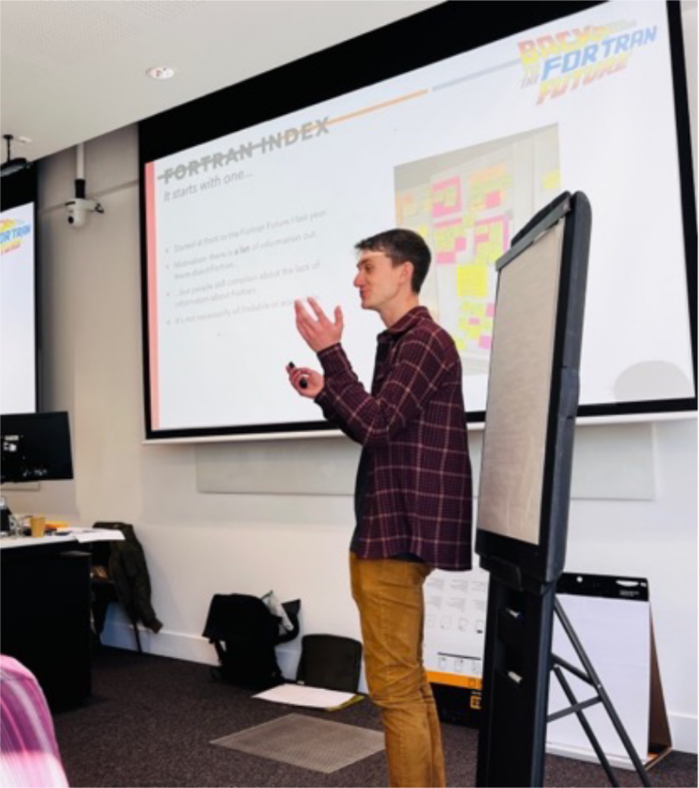 Young man (Joe) in red checkered shirt and brown trousers giving a talk in front of slides titled 'Fortran Index - It starts with one...'